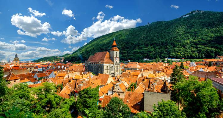 Terracotta rooftops of Brasov’s old town with the Black Church and a forested mountain backdrop under a bright sky.
