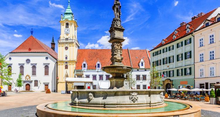 Sun-bathed historic main square with ornate fountain, pastel facades and outdoor cafés in Bratislava’s Old Town.
