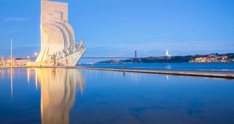 Monument to the Discoveries reflecting in still water with 25 de Abril Bridge at twilight.