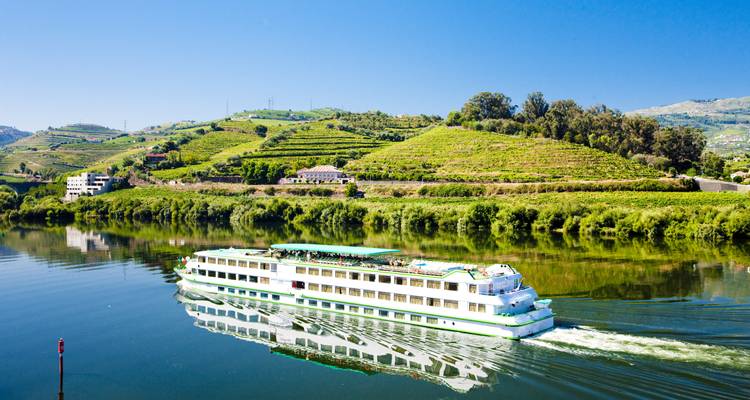 River cruise ship gliding along the mirror-calm Douro with terraced vineyards reflecting in the water on a bright sunny day.
