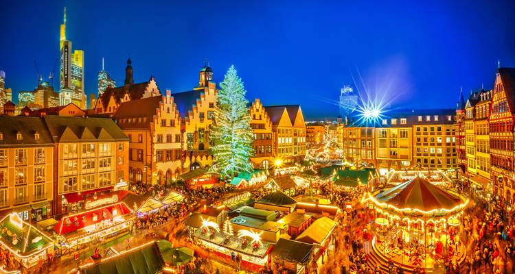 Panoramic night view over a bustling Christmas market with bright stalls, carousel, enormous tree and Frankfurt skyline beyond.