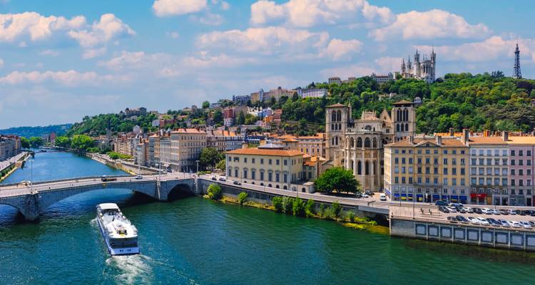 River scene with historic basilica on a hill and cruise boat passing beneath a bridge.
