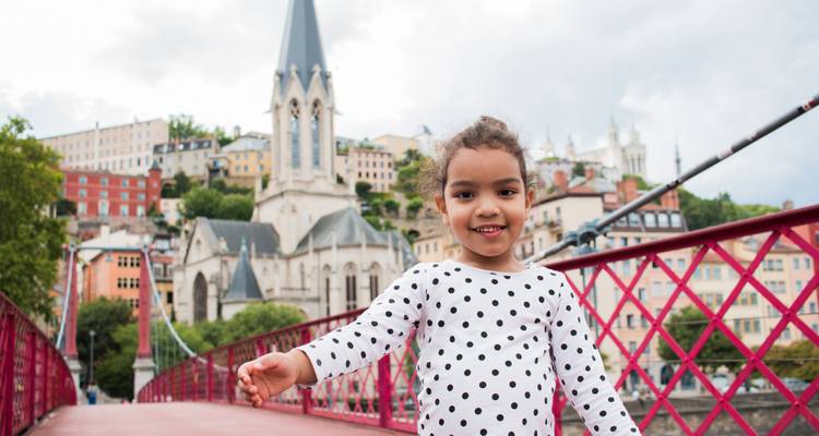 Smiling child on a red pedestrian bridge with historic hillside cityscape behind.