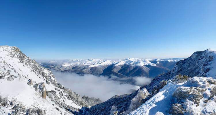 Panoramic snowy mountain vista with rugged cliffs framing a cloud-filled valley under a deep blue sky.