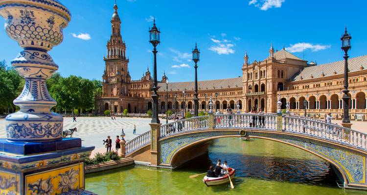 Escena brillante y soleada del puente de azulejos y el canal de la Plaza de España con turistas remando en botes bajo farolas ornamentadas.