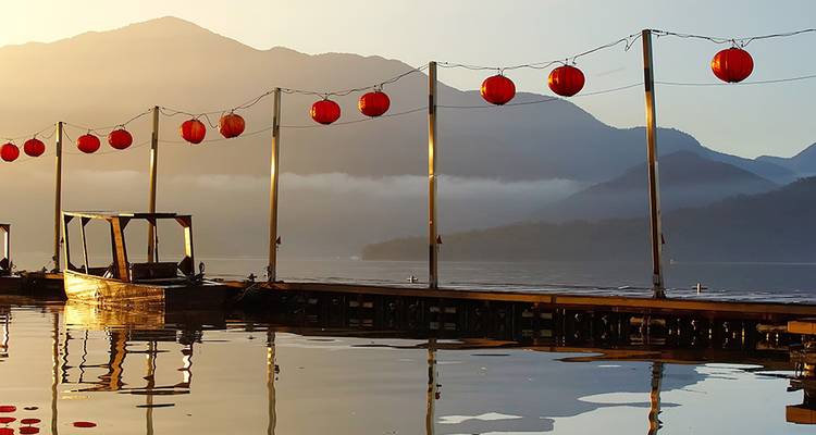 Rangée de lanternes rouges suspendues au-dessus d'un lac calme au coucher du soleil avec des montagnes brumeuses au-delà.