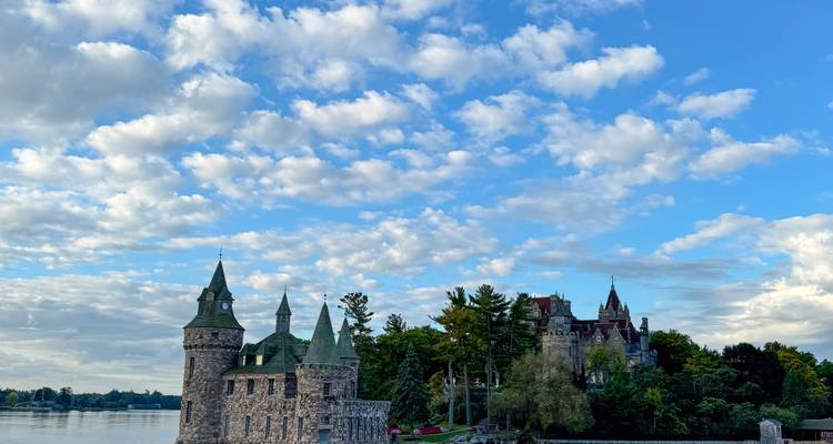 Fairy-tale stone castle with green towers sits on a wooded river island beneath a sky of scattered clouds.