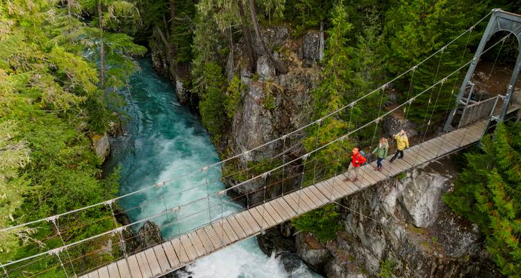 Three adventurers cross a narrow suspension bridge over a turquoise gorge in a lush forest.