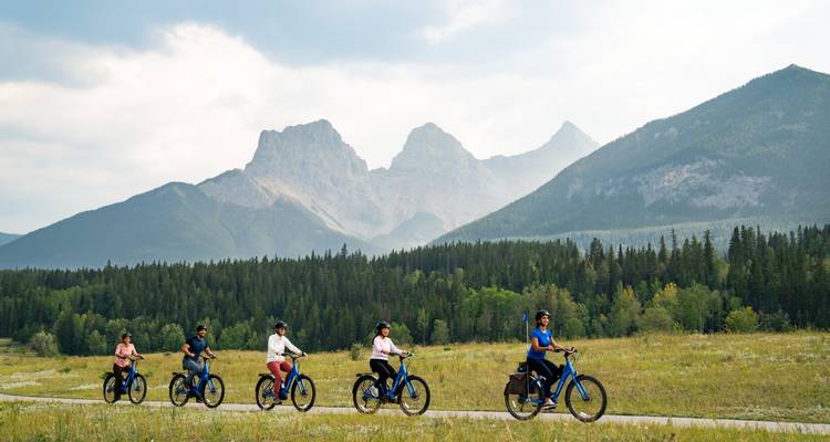 Five cyclists ride e-bikes across a grassy valley with jagged mountain peaks rising behind evergreen forests.