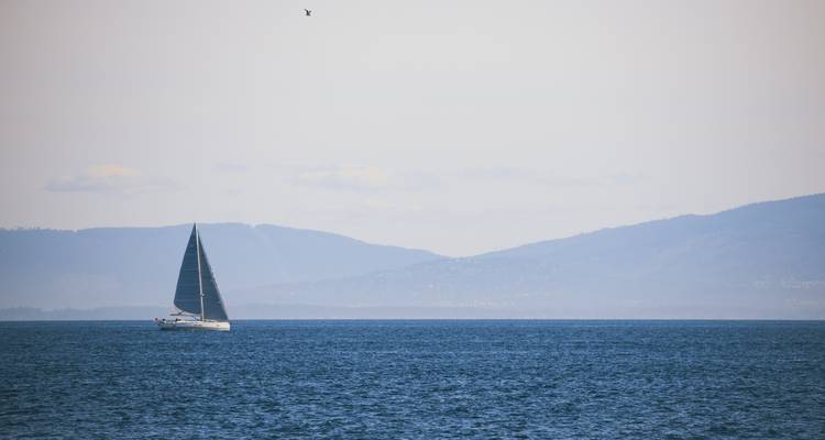 Un voilier solitaire glissant sur une mer bleu calme avec des montagnes voilées à l'horizon.