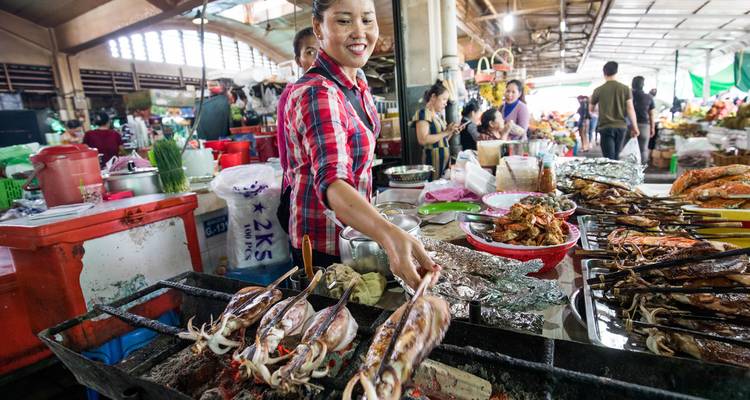 Un vendeur de marché souriant grille des brochettes de calmar frais sur des braises chaudes à l'intérieur d'un marché alimentaire cambodgien animé.