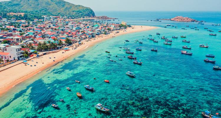 Aerial of a crescent beach and crystal-clear turquoise waters dotted with fishing boats near Quy Nhon.