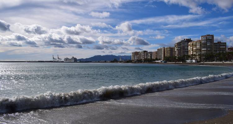 Gentle wave rolling onto Malaga city beach with skyline and distant mountains under partly cloudy skies.