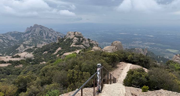 Sentier rocheux avec garde-corps menant à des vues panoramiques sur les montagnes de Montserrat.