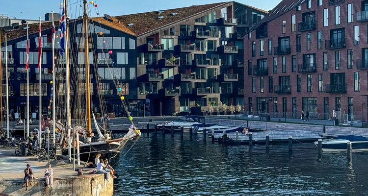 Moderne appartementen aan het kanaal met houten aanlegsteigers en afgemeerde zeilboten onder een helder lucht.
