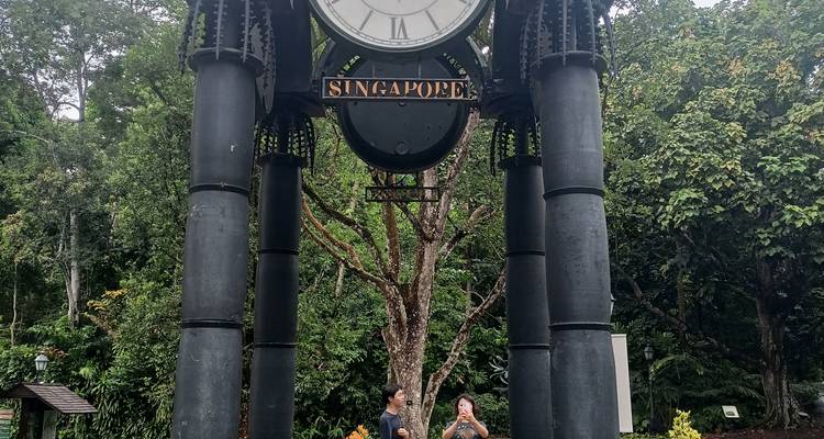 Hoher schwarzer gusseiserner Uhrenturm mit der Bezeichnung "Singapur" in einem grünen Park mit zwei Besuchern darunter
