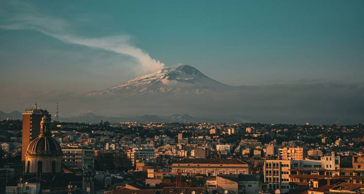 Le lointain Mont Etna aux pics enneigés fumant au-dessus de la silhouette de Catane baignée dans la lumière dorée du soir.