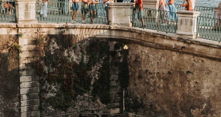 Pont piétonnier en pierre surplombant l'eau de mer avec des touristes appuyés contre la balustrade sous la lumière chaude du coucher de soleil.