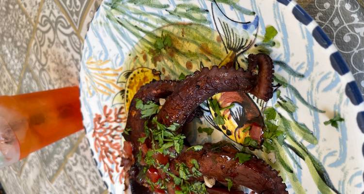 Gros plan sur une pieuvre grillée garnie d'herbes sur une assiette en céramique colorée à côté d'un cocktail à l'orange.
