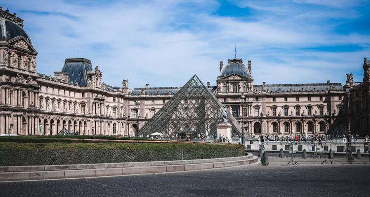 Perspective rapprochée de la Pyramide du Louvre encadrée par les ailes du palais historique sous un ciel dégagé.