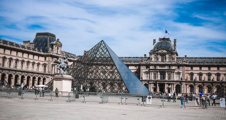 Vue générale de la vaste place de la Pyramide de verre du Louvre et du palais avec des visiteurs se promenant dans l'espace ouvert.