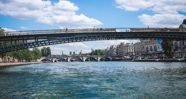 Vue de bas niveau du fleuve d'un pont piétonnier en fer enjambant la Seine avec des arches historiques au-delà.