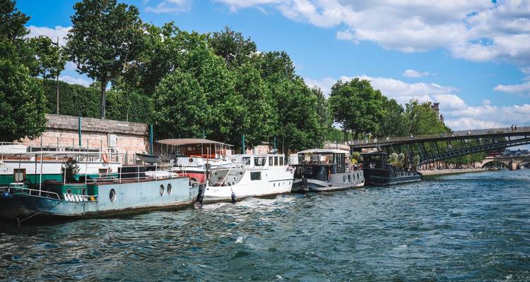 Rangée de péniches amarrées le long de la Seine sous des arbres feuillus avec un pont piétonnier en arrière-plan.