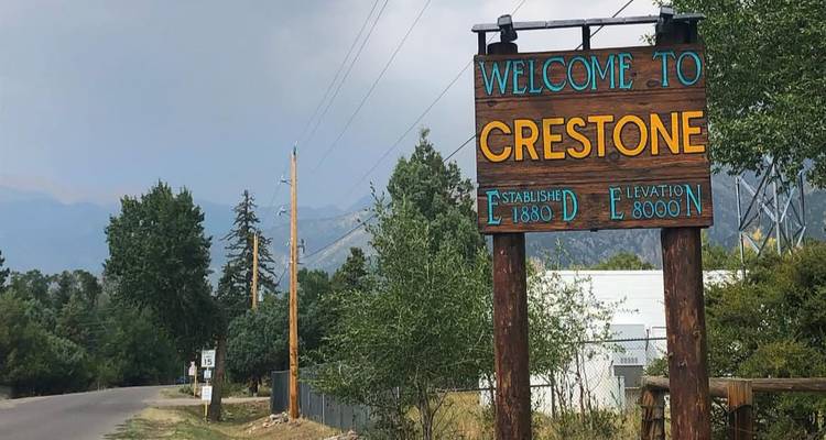 Rustic roadside sign reading 'Welcome to Crestone' with trees and distant mountains in the background.