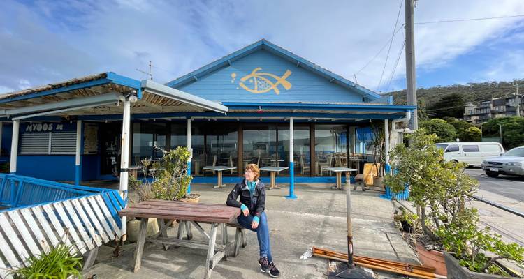 Personne assise à l'extérieur d'un café bord de mer bleu avec logo de poisson, avec des tables en plein air et une vue sur une ville côtière.