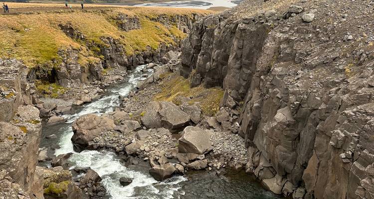 Gorge rocheuse profonde avec rivière à débit rapide et falaises couvertes de mousse, petits randonneurs visibles au-dessus.