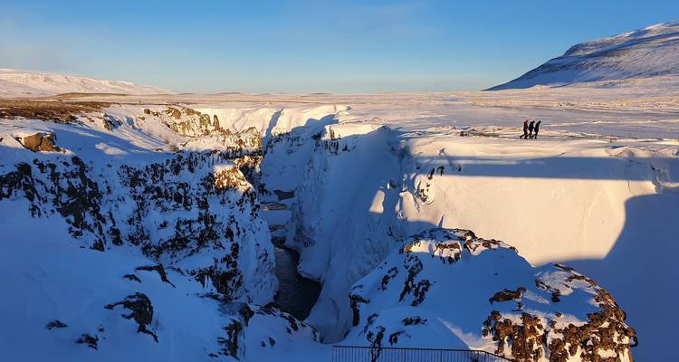 Gorge couverte de neige avec une rivière étroite traversant des falaises gelées sous un ciel d'hiver bleu clair ; quelques visiteurs se tiennent au bord projetant de longues ombres sur la surface blanche.