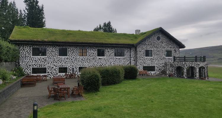 Maison d'hôtes islandaise traditionnelle avec toit de tourbe et façade distinctive en pierre de basalte noir et blanc se détachant sur un ciel nuageux et une pelouse verte avec du mobilier extérieur en bois.