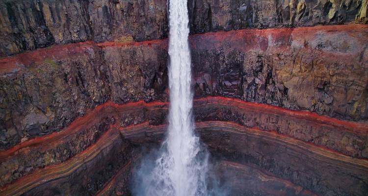 Une spectaculaire chute d'eau verticale dégringolant devant des couches de basalte rouge vif et brun à l'intérieur d'un profond canyon, capturée sous une perspective de face.