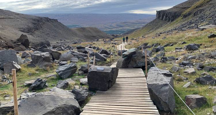 Passerelle en bois serpentant à travers un col montagneux rocheux avec des randonneurs au loin au milieu de rochers dispersés et de pentes herbeuses.