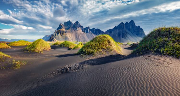 Des dunes de sable noir parsemées d'herbes vertes avec des montagnes aux contours dentelés qui s'élèvent en arrière-plan.