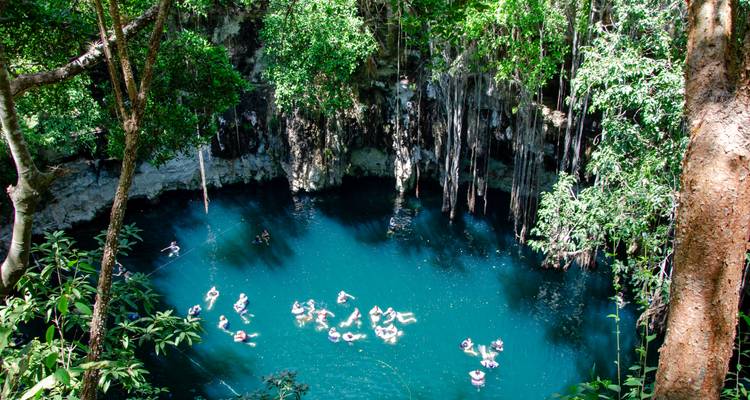 Luchtfoto van een diep blauw cenote omgeven door jungle-vegetatie met mensen die op het water drijven
