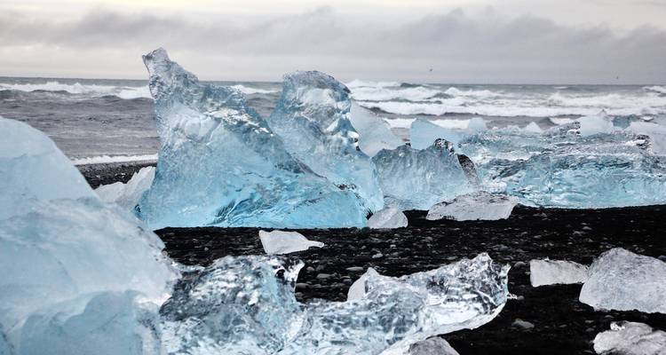 Large blue glacial ice chunks scattered across a black sand beach with stormy waves behind.