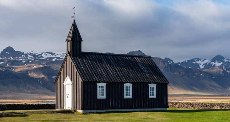 A small black wooden church standing alone on a grassy plain with rugged snow-dusted mountains behind.