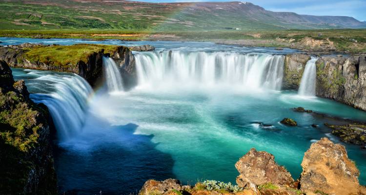 A powerful horseshoe-shaped waterfall plunging into a turquoise pool with a faint rainbow arcing above.