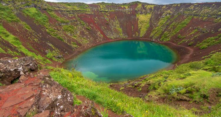 A vivid blue crater lake surrounded by red volcanic rock walls and bright green moss.