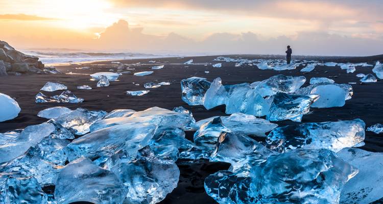 Glittering ice blocks on black sand at sunset with a lone figure gazing toward the ocean.