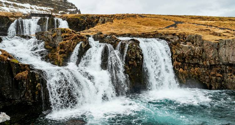 Tiered waterfalls tumbling over rocky cliffs into a bright blue pool amid golden tundra.