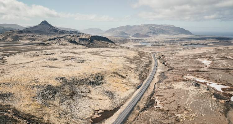 Vue aérienne par drone d'une route solitaire serpentant à travers une péninsule volcanique aride avec des montagnes voilées au loin.