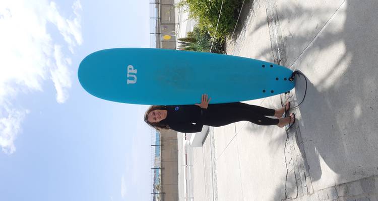 Femme en combinaison de plongée souriante tenant une planche de surf turquoise portant l'étiquette « UP » sur un front de plage pavé ensoleillé.