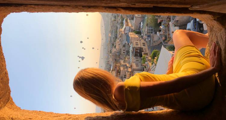 Un voyageur vêtu d'une robe jaune est assis sur un rebord de pierre encadré par une fenêtre en arc avec des ballons s'élevant au-dessus de Göreme.