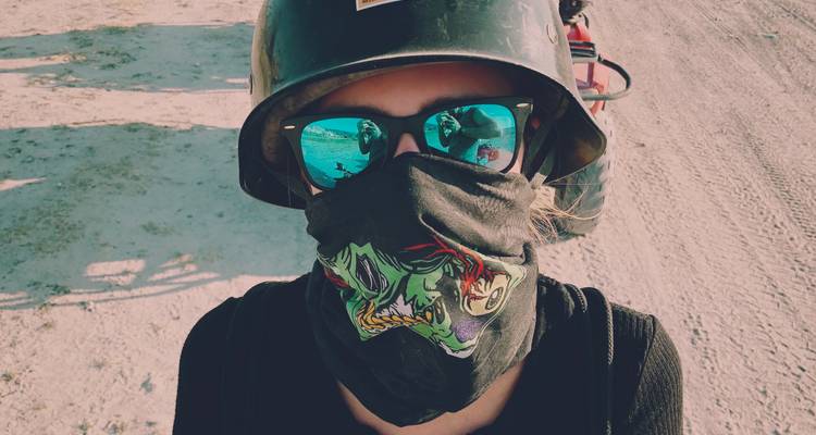 Un voyageur aventurier portant un casque, des lunettes de soleil et un bandana à motifs se tient sur un sentier poussiéreux.