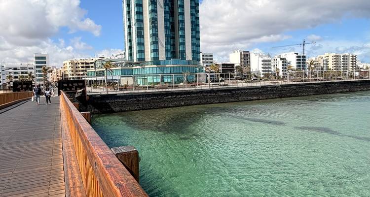 Une promenade en bois menant sur une eau turquoise vers un hôtel moderne de grande hauteur sur le front de mer d'Arrecife.