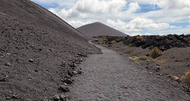 Un sendero de ceniza negra que conduce hacia un volcán en forma de cono bajo nubes dispersas.