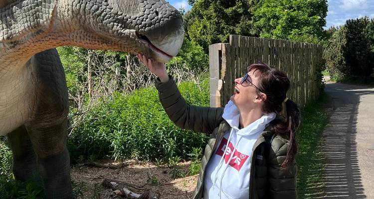 Femme souriante tendant le bras pour toucher le museau d'une grande maquette de dinosaure dans une exposition extérieure verdoyante