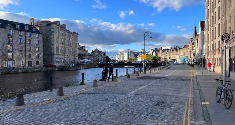 Promenade riveraine pavée à Leith avec des bâtiments historiques, des réverbères et des piétons marchant sous un ciel bleu éclatant